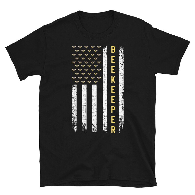 Pode incluir: Camiseta preta com um design de bandeira americana branca e dourada e a palavra "BEEKEEPER" verticalmente do lado direito da bandeira.