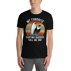 Może przedstawiać: Czarny T-shirt z grafiką retro zachodu słońca i dwoma deskami surfingowymi. Tekst brzmi "Moi ulubieni surferzy nazywają mnie tatą."
