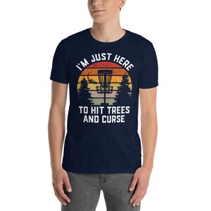 May include: A navy blue t-shirt with a vintage-style sunset graphic featuring a disc golf basket and trees. The text "I'm Just Here To Hit Trees And Curse" is printed in white on the shirt.