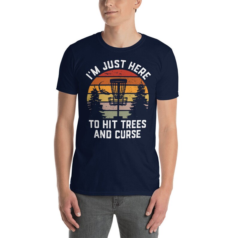 May include: A navy blue t-shirt with a vintage-style sunset graphic featuring a disc golf basket and trees. The text "I'm Just Here To Hit Trees And Curse" is printed in white on the shirt.