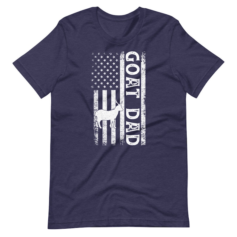 以下が含まれることがあります： アメリカ国旗のデザインが施されたネイビーブルーのTシャツ。旗には白いヤギのシルエットと白い文字で「GOAT DAD」と書かれています。