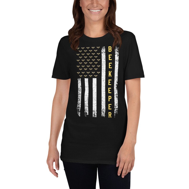 Pode incluir: Camiseta preta com um design de bandeira americana branca e dourada. A bandeira tem uma fileira de abelhas no topo e a palavra "BEEKEEPER" verticalmente para baixo no lado direito.