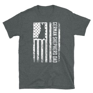 May include: A dark heather grey t-shirt with a white silhouette of a German Shepherd dog in front of an American flag. The text "GERMAN SHEPHERD DAD" is printed on the right side of the flag.