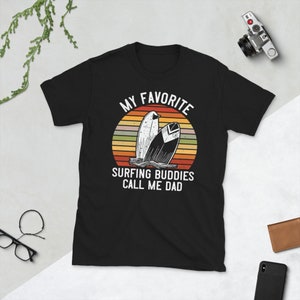 Może przedstawiać: Czarny T-shirt z grafiką retro zachodu słońca i dwoma deskami surfingowymi. Na koszulce nadrukowany jest tekst "My Favorite Surfing Buddies Call Me Dad".