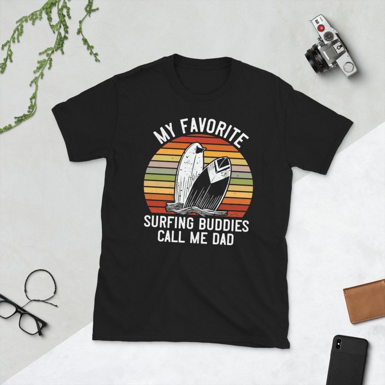 Może przedstawiać: Czarny T-shirt z grafiką retro zachodu słońca i dwoma deskami surfingowymi. Na koszulce nadrukowany jest tekst "My Favorite Surfing Buddies Call Me Dad".