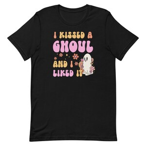 Op de afbeelding: Zwart T-shirt met een roze en oranje afbeelding met de tekst "I kissed a ghoul and I liked it". De afbeelding bevat ook een wit spook met roze bloemen.