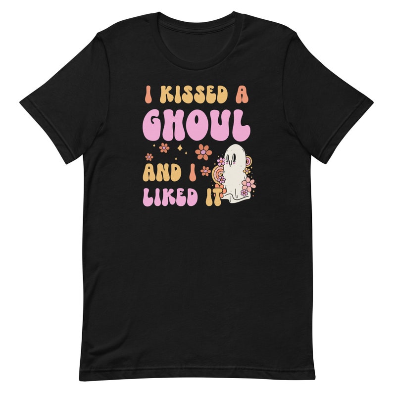 Op de afbeelding: Zwart T-shirt met een roze en oranje afbeelding met de tekst "I kissed a ghoul and I liked it". De afbeelding bevat ook een wit spook met roze bloemen.