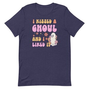 Op de afbeelding: Een marineblauw T-shirt met een roze en oranje afbeelding waarop staat "I kissed a ghoul and I liked it". De afbeelding toont een spook met een roze bloem erachter.
