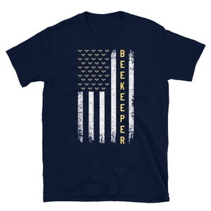 Pode incluir: Uma camiseta azul marinho com um design de bandeira americana branca e dourada. A bandeira tem uma fileira de abelhas no topo e a palavra "BEEKEEPER" verticalmente para baixo no lado direito.