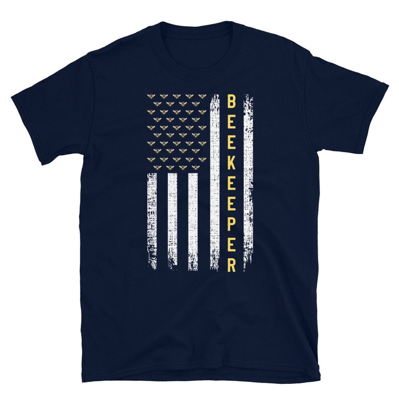 Pode incluir: Uma camiseta azul marinho com um design de bandeira americana branca e dourada. A bandeira tem uma fileira de abelhas no topo e a palavra "BEEKEEPER" verticalmente para baixo no lado direito.