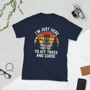 May include: A navy blue t-shirt with a vintage-style graphic of a disc golf basket in front of a sunset. The text reads "I'm Just Here To Hit Trees And Curse".