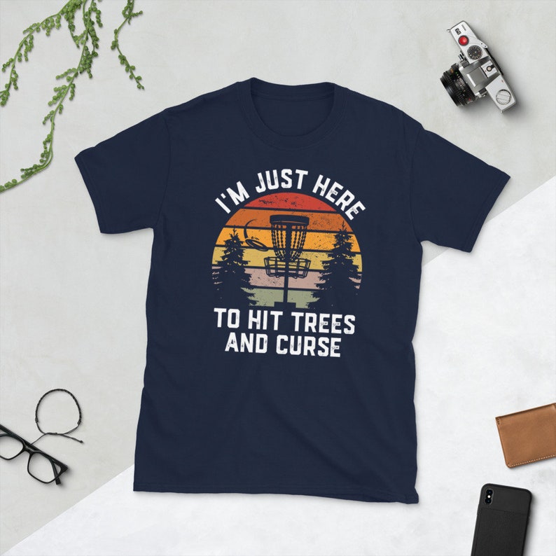May include: A navy blue t-shirt with a vintage-style graphic of a disc golf basket in front of a sunset. The text reads "I'm Just Here To Hit Trees And Curse".