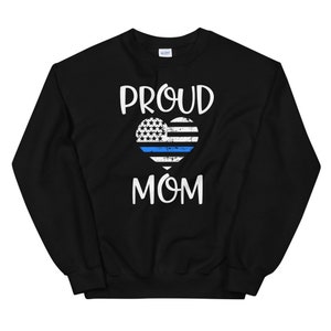May include: Black sweatshirt with the words "PROUD MOM" in white script. A heart-shaped graphic features a blue line, white stripes, and a field of white stars, resembling a flag. The sweatshirt is made of a soft material.