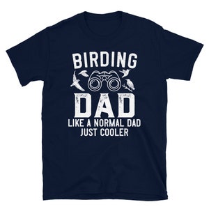Op de afbeelding: Donkerblauw T-shirt met witte tekst die "Birding Dad Like a Normal Dad Just Cooler" luidt. Het shirt heeft een verrekijker en een vogel illustratie.