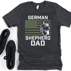 K&ouml;nnte beinhalten: Ein dunkelgraues T-Shirt mit einem wei&szlig;en Grafikdesign. Die Grafik zeigt den Kopf eines Deutschen Sch&auml;ferhundes vor einer amerikanischen Flagge mit gr&uuml;nen und wei&szlig;en Streifen. Der Text "GERMAN SHEPHERD DAD" ist unter dem Bild gedruckt.