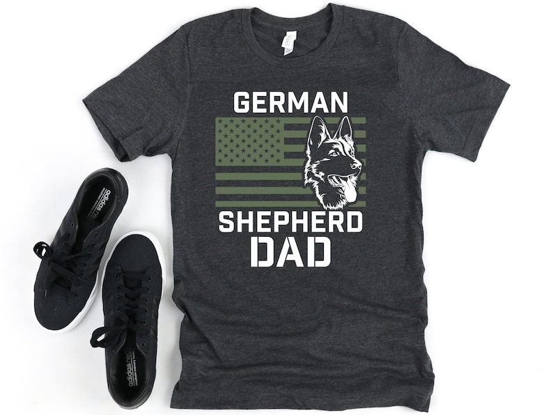 K&ouml;nnte beinhalten: Ein dunkelgraues T-Shirt mit einem wei&szlig;en Grafikdesign. Die Grafik zeigt den Kopf eines Deutschen Sch&auml;ferhundes vor einer amerikanischen Flagge mit gr&uuml;nen und wei&szlig;en Streifen. Der Text "GERMAN SHEPHERD DAD" ist unter dem Bild gedruckt.