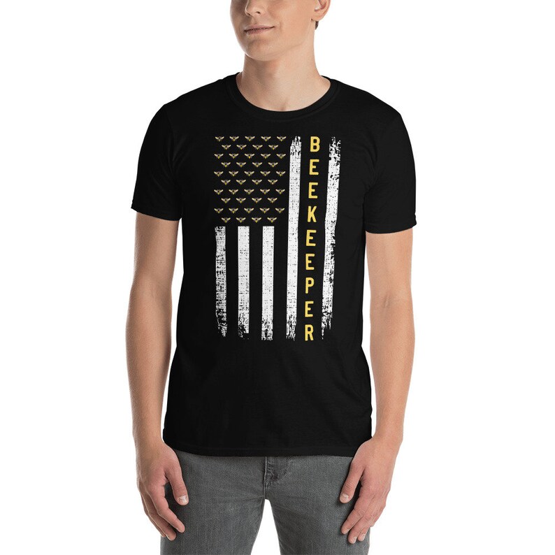 Pode incluir: Camiseta preta com um design de bandeira americana branca e amarela. A bandeira tem um visual desgastado e a palavra "BEEKEEPER" est&aacute; escrita verticalmente do lado direito da bandeira.