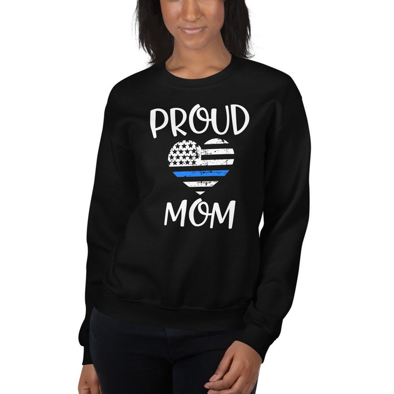 May include: Black sweatshirt with the words "PROUD MOM" in white. A heart-shaped design features the American flag with a blue line. The sweatshirt is a crew neck style.