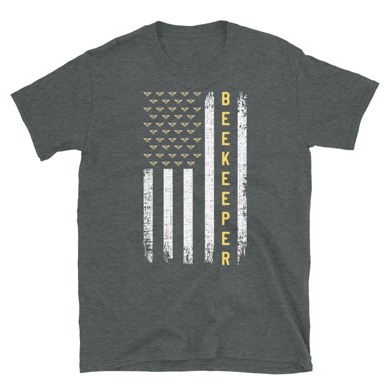Pode incluir: Uma camiseta cinza escura mesclada com um design de bandeira americana branca e amarela. A bandeira tem uma fileira de abelhas no topo e a palavra "BEEKEEPER" verticalmente do lado direito.