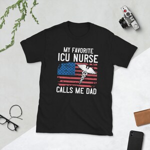 May include: Black t-shirt with a white graphic that says "My Favorite ICU Nurse Calls Me Dad". The graphic also includes an American flag with a medical caduceus symbol.