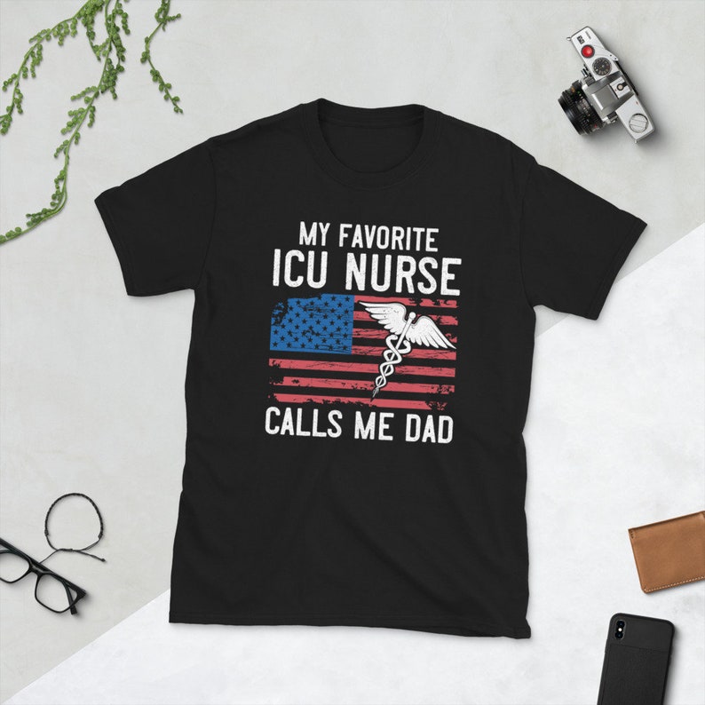 May include: Black t-shirt with a white graphic that says "My Favorite ICU Nurse Calls Me Dad". The graphic also includes an American flag with a medical caduceus symbol.