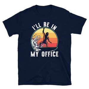 May include: A navy blue t-shirt with a retro-style graphic of a surfer riding a wave. The text "I'll be in my office" is printed in white on the shirt.