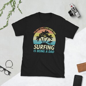 Pu&ograve; includere: T-shirt nero con una grafica di tramonto retr&ograve; e palme. Il testo recita "L'unica cosa che amo pi&ugrave; del surf &egrave; essere pap&agrave;."