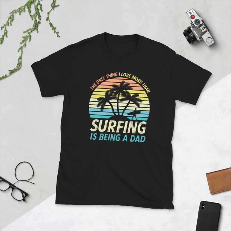 Pu&ograve; includere: T-shirt nero con una grafica di tramonto retr&ograve; e palme. Il testo recita "L'unica cosa che amo pi&ugrave; del surf &egrave; essere pap&agrave;."