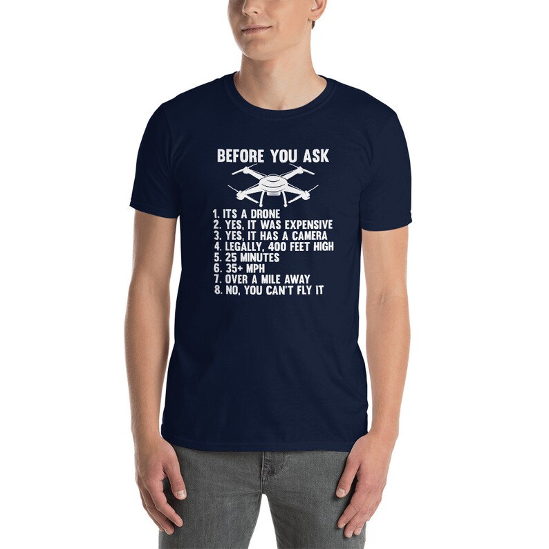 May include: A navy blue t-shirt with white text that reads "BEFORE YOU ASK" and lists the features of a drone. The text includes: "ITS A DRONE", "YES, IT WAS EXPENSIVE", "YES, IT HAS A CAMERA", "LEGALLY, 400 FEET HIGH", "25 MINUTES", "35+ MPH", "OVER A MILE AWAY", and "NO, YOU CAN'T FLY IT".