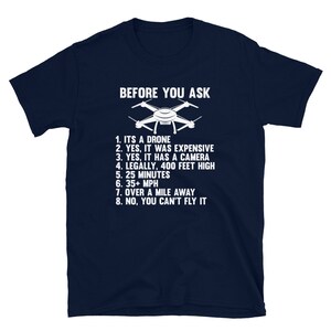 May include: A navy blue t-shirt with white text that reads "BEFORE YOU ASK" and lists eight bullet points about a drone. The text includes the following: "ITS A DRONE", "YES, IT WAS EXPENSIVE", "YES, IT HAS A CAMERA", "LEGALLY, 400 FEET HIGH", "25 MINUTES", "35+ MPH", "OVER A MILE AWAY", and "NO, YOU CAN'T FLY IT".