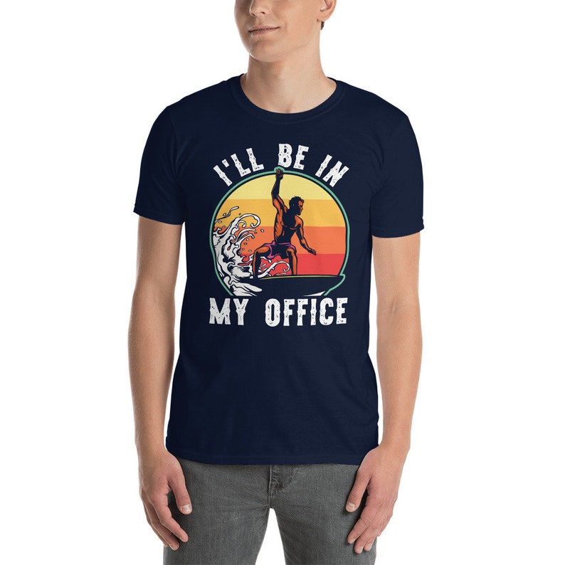 May include: A navy blue t-shirt with a vintage-style graphic of a surfer riding a wave. The graphic includes a sunset and the text "I'll be in my office".