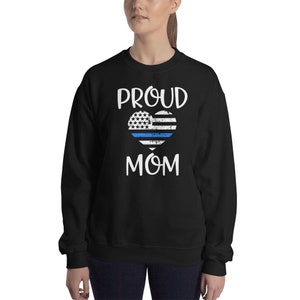 May include: Black sweatshirt with the words "PROUD MOM" in white. A heart-shaped design features a blue line, stars, and stripes, resembling a flag. The sweatshirt is a casual garment.