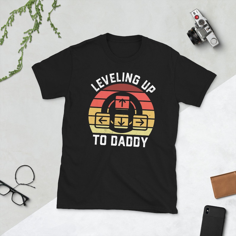 May include: Black t-shirt with a retro-style graphic that says "Leveling Up To Daddy" with an arrow pointing up and left, right, and down arrows around it.