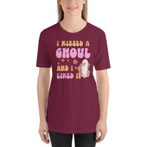 Op de afbeelding: Een maroon t-shirt met een retro-stijl grafische afbeelding met de tekst "I kissed a ghoul and I liked it". De afbeelding toont een roze geest met bloemen en sterren.