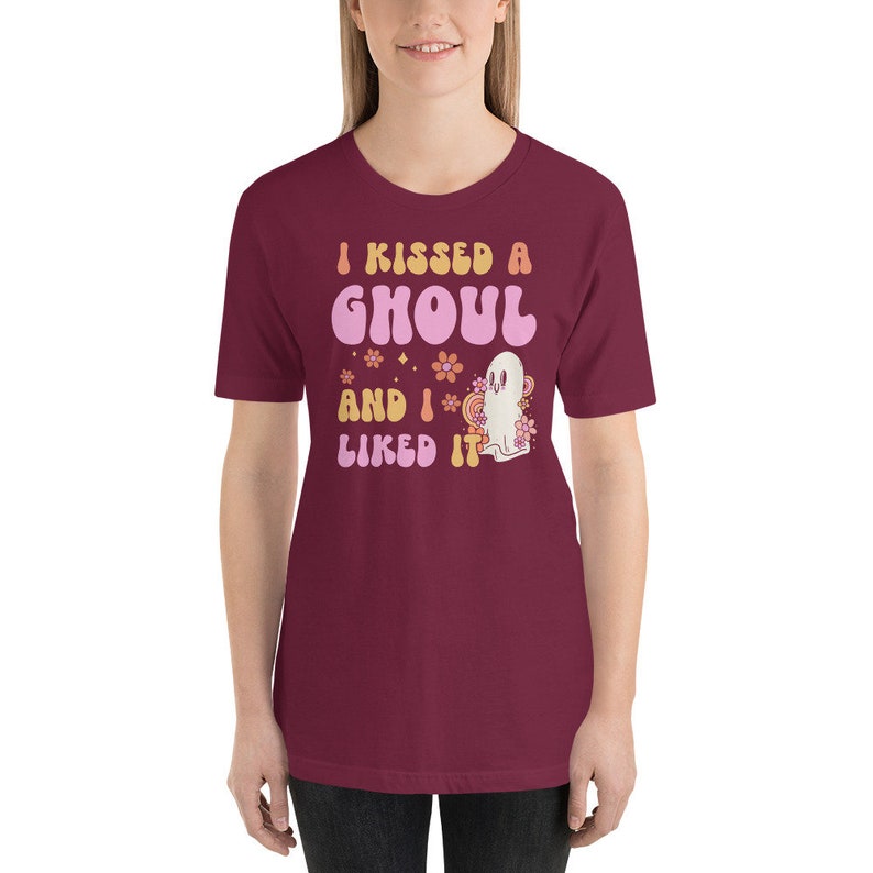 Op de afbeelding: Een maroon t-shirt met een retro-stijl grafische afbeelding met de tekst "I kissed a ghoul and I liked it". De afbeelding toont een roze geest met bloemen en sterren.