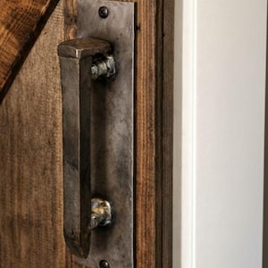 May include: A rustic, metal door pull with a rectangular backplate and a curved handle. The handle is attached with bolts to a dark, stained wooden door. The door is made of vertical wooden planks. The door is next to a white door frame.