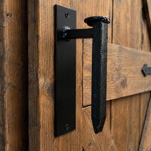 May include: A black metal door handle featuring a rectangular mounting plate secured with two screws and a vertical, railroad spike-style grip. The handle is mounted onto a brown wooden door with visible grain and diagonal bracing patterns.