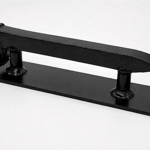 May include: A black metal door pull with a rectangular base and a horizontal, railroad spike-shaped handle. The handle is mounted on two cylindrical supports. The door pull is designed for a rustic or industrial aesthetic.