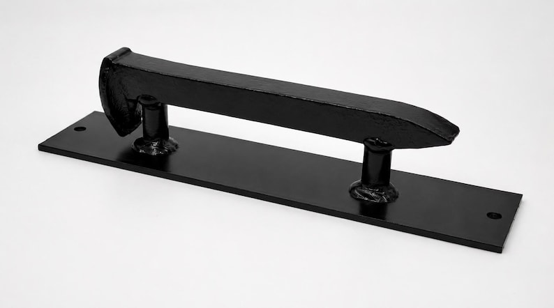 May include: A black metal door pull with a rectangular base and a horizontal, railroad spike-shaped handle. The handle is mounted on two cylindrical supports. The door pull is designed for a rustic or industrial aesthetic.