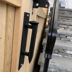 May include: Black metal gate handle and hardware attached to a wooden gate. The handle is a stylized design with a vertical plate and a horizontal bar. The gate is made of light-colored wood. A black metal railing is visible in the background.