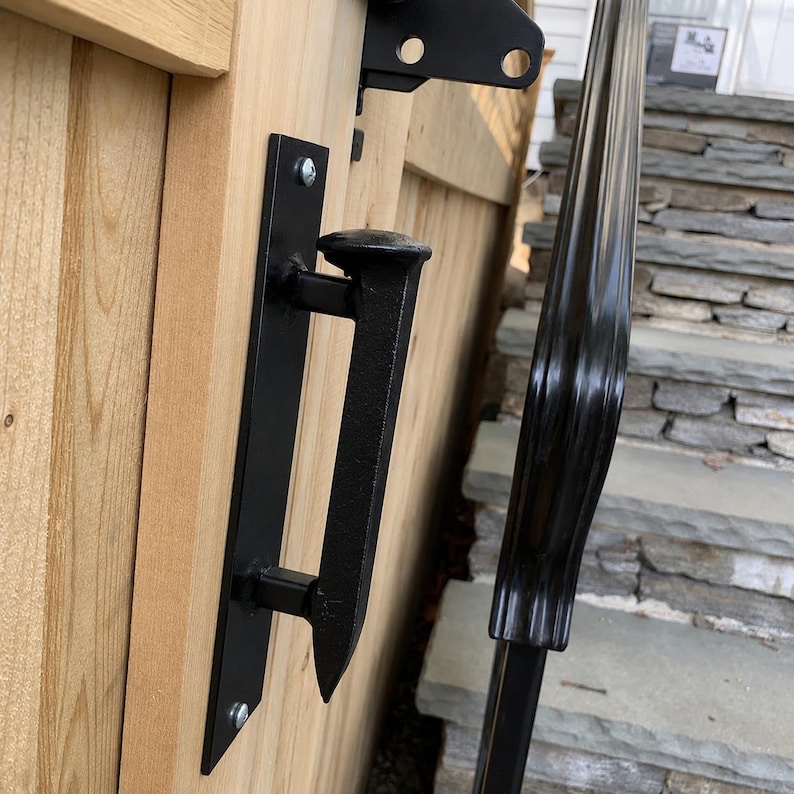 May include: Black metal gate handle and hardware attached to a wooden gate. The handle is a stylized design with a vertical plate and a horizontal bar. The gate is made of light-colored wood. A black metal railing is visible in the background.