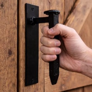 May include: Black iron door handle with a rectangular backplate and a textured, nail-shaped grip. The handle is mounted on a wooden door with visible wood grain.