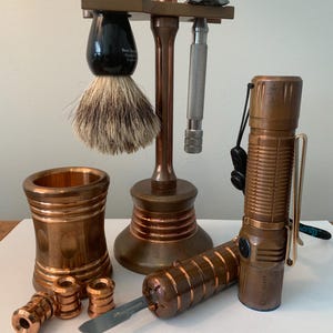 May include: A collection of grooming accessories and tools in a copper finish. Includes a shaving brush, razor, stand, small containers, a flashlight, and a utility knife. The items are arranged on a white surface.