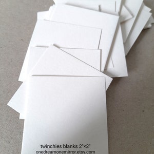 May include: A stack of blank, white square cards, each measuring 2" x 2". The cards are arranged in a slightly fanned-out pattern. Text on one card reads "twinchies blanks 2"x2" onedreamonemirror.etsy.com".