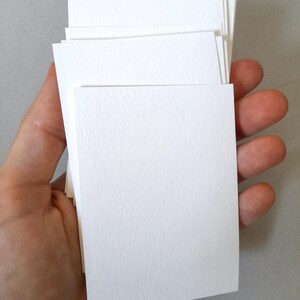 May include: A stack of blank, white rectangular cards held in a hand. The cards are stacked neatly, with a slightly textured surface. The edges of the cards are clean and crisp, suggesting a high-quality paper stock. The background is a neutral gray.
