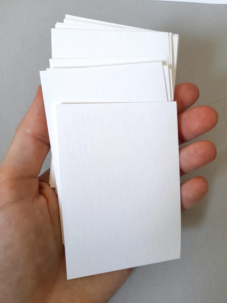 May include: A stack of blank, white rectangular cards held in a hand. The cards are stacked neatly, with a slightly textured surface. The edges of the cards are clean and crisp, suggesting a high-quality paper stock. The background is a neutral gray.