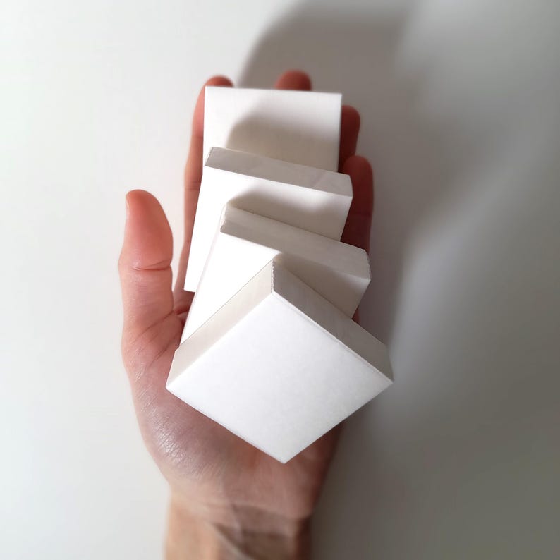 May include: A hand holding a stack of white, square, paper-like blocks. The blocks are arranged in a staggered, cascading pattern, with each block slightly offset from the one below. The background is a plain white surface.