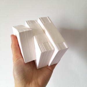 May include: A hand holding three stacks of white paper. Each stack is rectangular, with a clean, crisp edge. The paper appears to be blank, with a smooth surface. The background is a plain white wall.