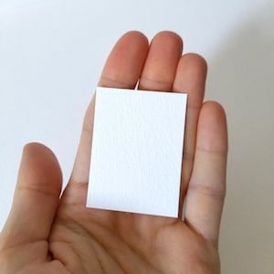 May include: A small, rectangular, white piece of paper with a textured surface is held in an open hand. The paper is centered in the frame, and the hand is slightly out of focus.