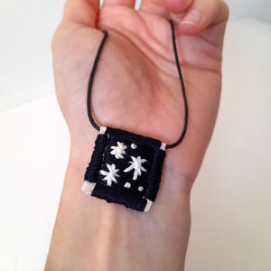 Op de afbeelding: Een handgemaakte ketting met een donkerblauwe vierkante hanger met witte geborduurde sterrenpatronen. De hanger is bevestigd aan een zwart koord. De ketting wordt in een hand gehouden.